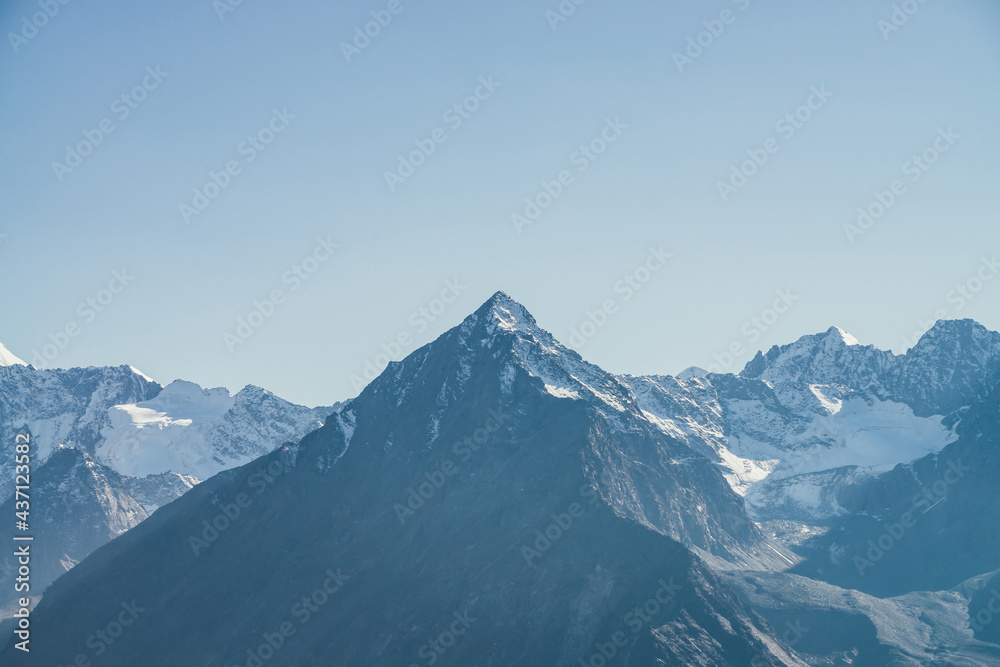 Atmospheric mountain landscape with black mountain silhouette with sharp rocky pinnacle with snow in sunlight. Alpine scenery with dark snow-covered mountain with peaked top in sunshine under blue sky