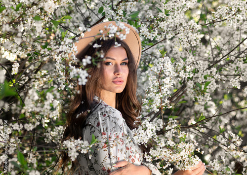 Natural beauty brunette woman in a hat with bouquet of cherry branches walks in the summer garden, enjoying the blooming spring nature