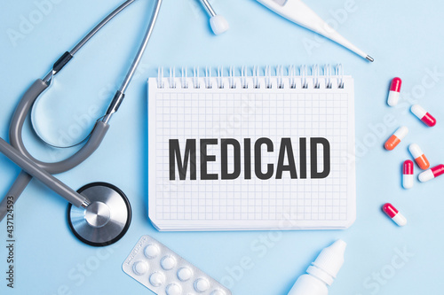 The word medicaid written on a white notepad on a blue background near a stethoscope, syringe, electronic thermometer and pills. Medical concept photo