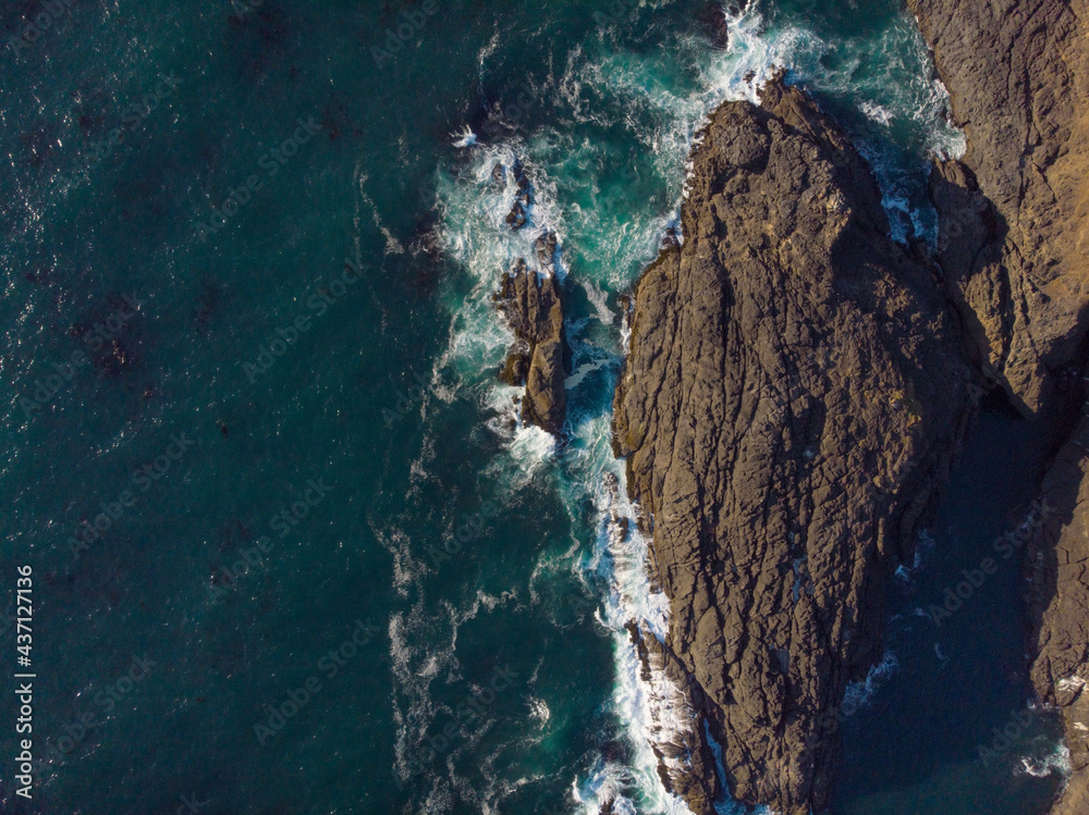 In the photo we see a seascape. The deep turquoise ocean water and dark brown rocks create a contrast. Bright lighting. No people. View from above. Aerial photography. Background. Wallpaper. Texture