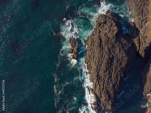 In the photo we see a seascape. The deep turquoise ocean water and dark brown rocks create a contrast. Bright lighting. No people. View from above. Aerial photography. Background. Wallpaper. Texture