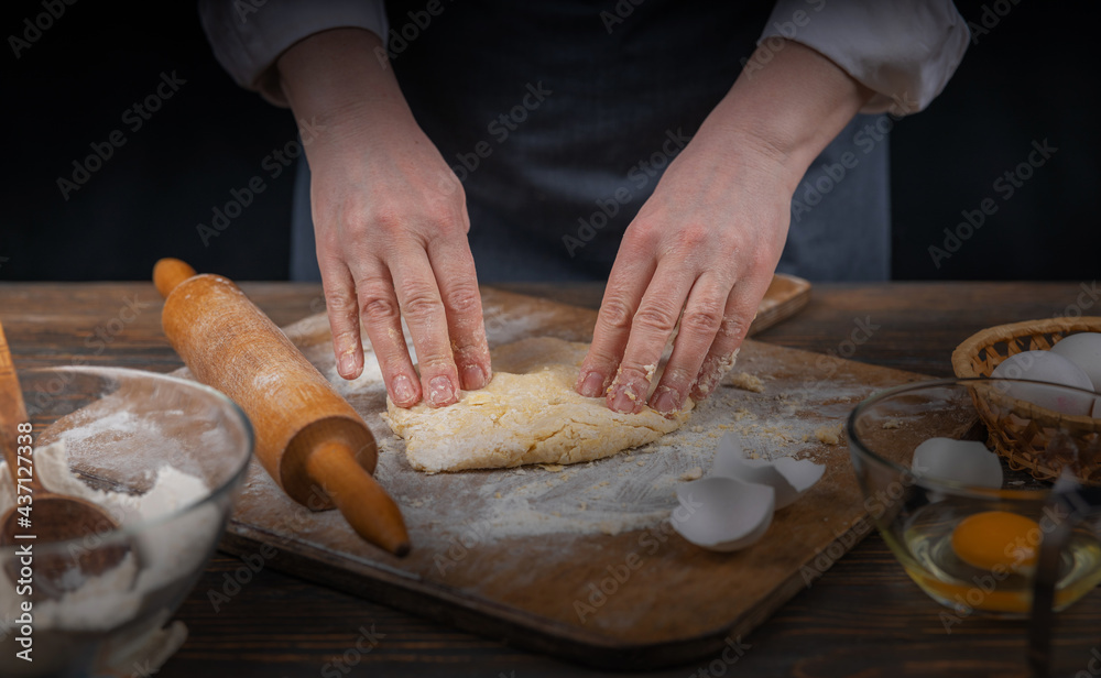 Women's hands, flour and dough. A woman in an apron cooking dough for homemade baking, a rustic home cozy atmosphere, a dark background with unusual lighting. Women's hands, flour and dough. A woman in an apron cooking dough for homemade baking, a rustic home cozy atmosphere, a dark background with unusual lighting.