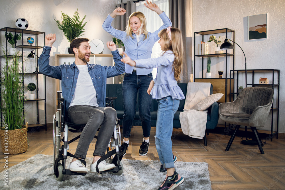 Handsome man in wheelchair dancing with lovely wife and pretty daughter at home. Rehabilitation and support concept. Family time.