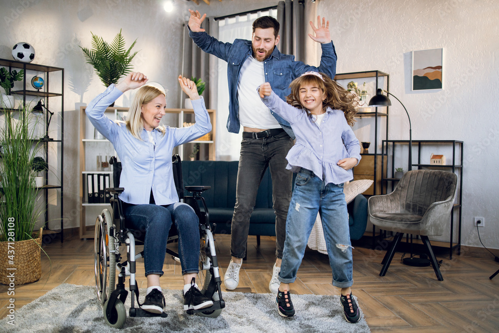 Happy woman in wheelchair dancing with beloved husband and pretty daughter at living room. Family spending actively time together. People with disabilities.