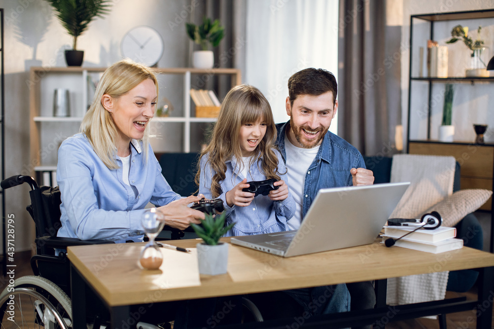 Disabled woman playing video games at home with her pretty daughter and handsome husband. Cheeful parents and kid using modern laptop and joysticks for entertainment. Disabled woman playing video games at home with her pretty daughter and handsome husband. Cheeful parents and kid using modern laptop and joysticks for entertainment.