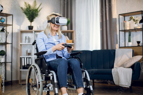 Smiling woman with blond hair sitting in wheelchair and playing virtual games with joystick and 3D glasses. Recovery process at home with entertainment.
