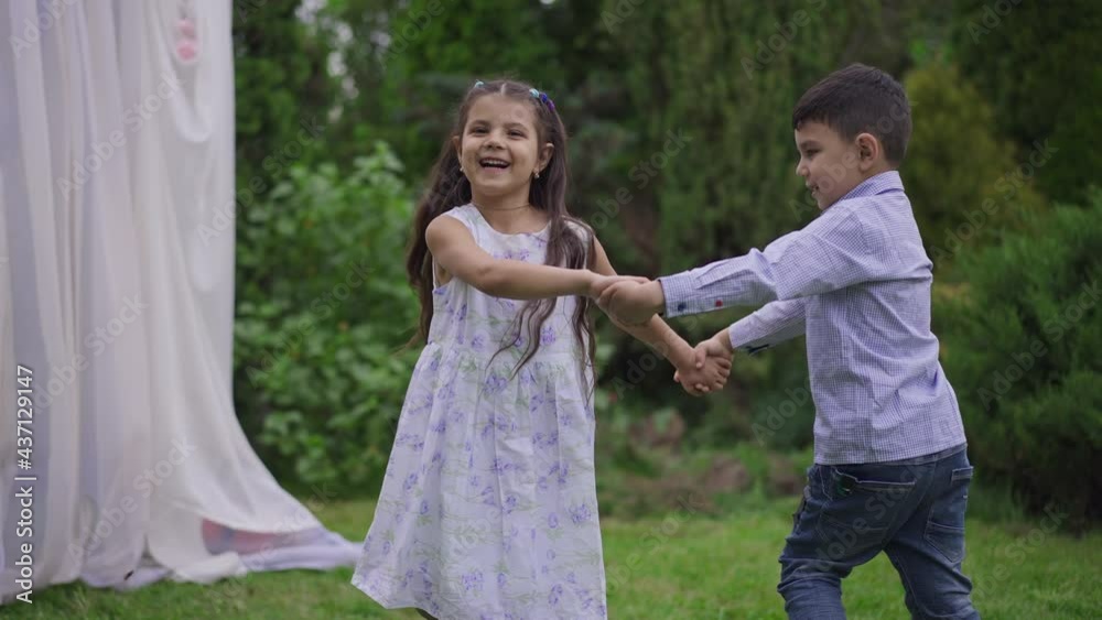 Zoom in to cheerful Middle Eastern little boy and girl spinning jumping holding hands at wedding altar in summer garden. Happy joyful children enjoying marriage ceremony outdoors having fun Zoom in to cheerful Middle Eastern little boy and girl spinning jumping holding hands at wedding altar in summer garden. Happy joyful children enjoying marriage ceremony outdoors having fun