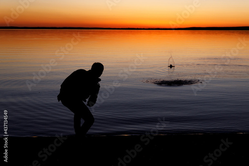 Fotografija Silhouette of a man throws stones into the lake at sunset