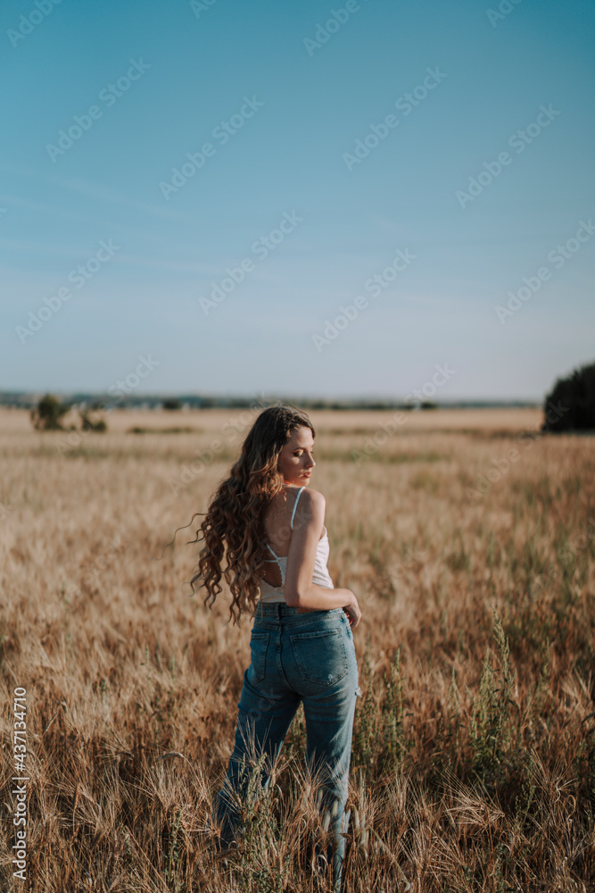 Chica joven atractiva posando en mitad de un campo de trigo al atardecer con tonos dorados Chica joven atractiva posando en mitad de un campo de trigo al atardecer con tonos dorados