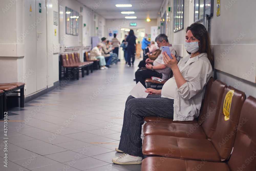 Pregnant woman with phone in clinic lobby with medical mask on face Pregnant woman with phone in clinic lobby with medical mask on face