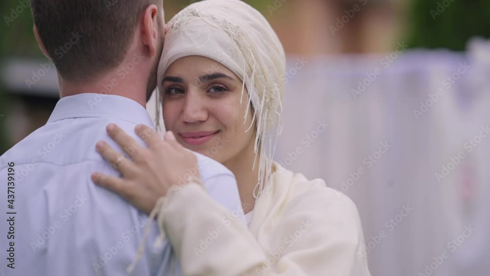 Portrait of happy charming Middle Eastern bride in hijab hugging unrecognizable groom looking at camera smiling. Loving affectionate woman embracing man on marriage ceremony outdoors Portrait of happy charming Middle Eastern bride in hijab hugging unrecognizable groom looking at camera smiling. Loving affectionate woman embracing man on marriage ceremony outdoors