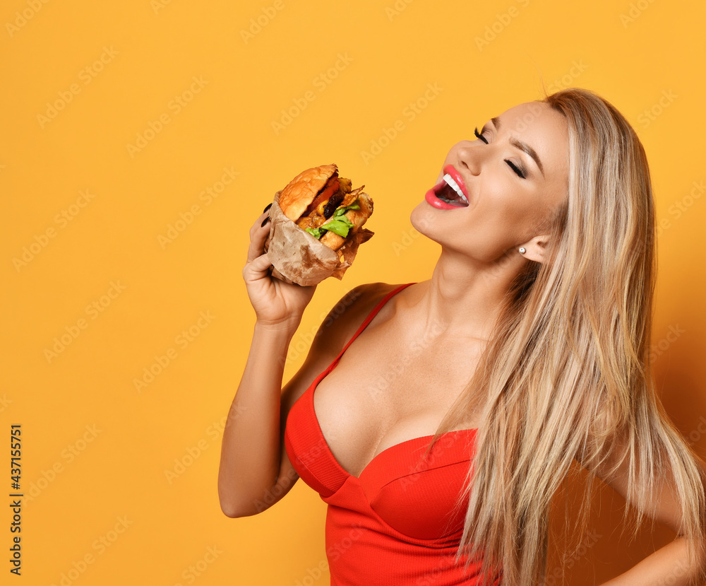 Young beautiful sexy smiling blonde woman in red swimsuit with deep neckline holding fresh hamburger in hands over yellow wall background. Diet, slimming, fast food, weight loss concept