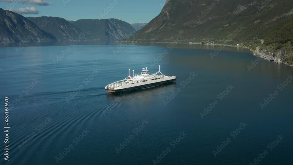 Kaupanger-Laerdal ferry crossing. An electric ferry moving fast on the still waters of the fjord nearing the harbour. Mountains, covered with pine forest rise from the water. The sky is clear.