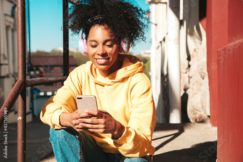Beautiful black woman with afro curls hairstyle.Smiling model in yellow hoodie.Sexy carefree female enjoying listening music in wireless headphones.Posing on street background at sunset.Holds phone