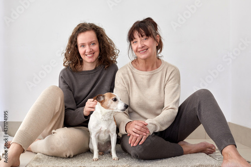 Cheerful adult daughter and middle aged mother sitting on floor with cute dog at home