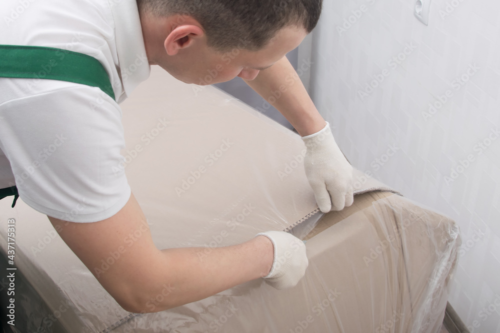 Obraz premium the delivery man in protective gloves, cuts the dense polyethylene, to protect the cardboard from moisture, close-up