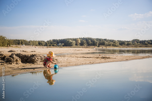 Little cute 2 year caucasian girl in red swimsuit and panama sitting on coastline and playing with bucket and scoop. Nature landscape. Copy space. Calm water reflection. Evening summer time. Traveler