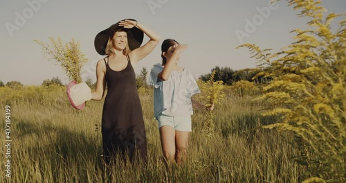 Mother and daughter, walk on a yellow summer lawn under the sun, non urban scene. Girls going straight ahead slowly, looking at horizon. Holding hat in a hand. Nature background, weekend out of city