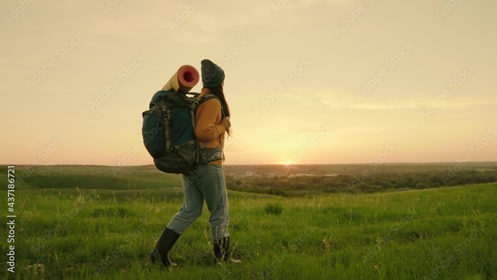 Free woman traveler walking through the meadow in summer. Girl traveler travels along top of green hill in sun, meditation, ecotourism, hike. An active young caucasian woman with backpack goes on hike