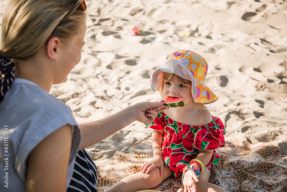 A beautiful young mother and little 2-year daughter are sitting on sand and eating a watermelon. Picnic on the beach. Sea travel holiday. A family care. Happiness lifestyle. Love. Close-up portrait A beautiful young mother and little 2-year daughter are sitting on sand and eating a watermelon. Picnic on the beach. Sea travel holiday. A family care. Happiness lifestyle. Love. Close-up portrait