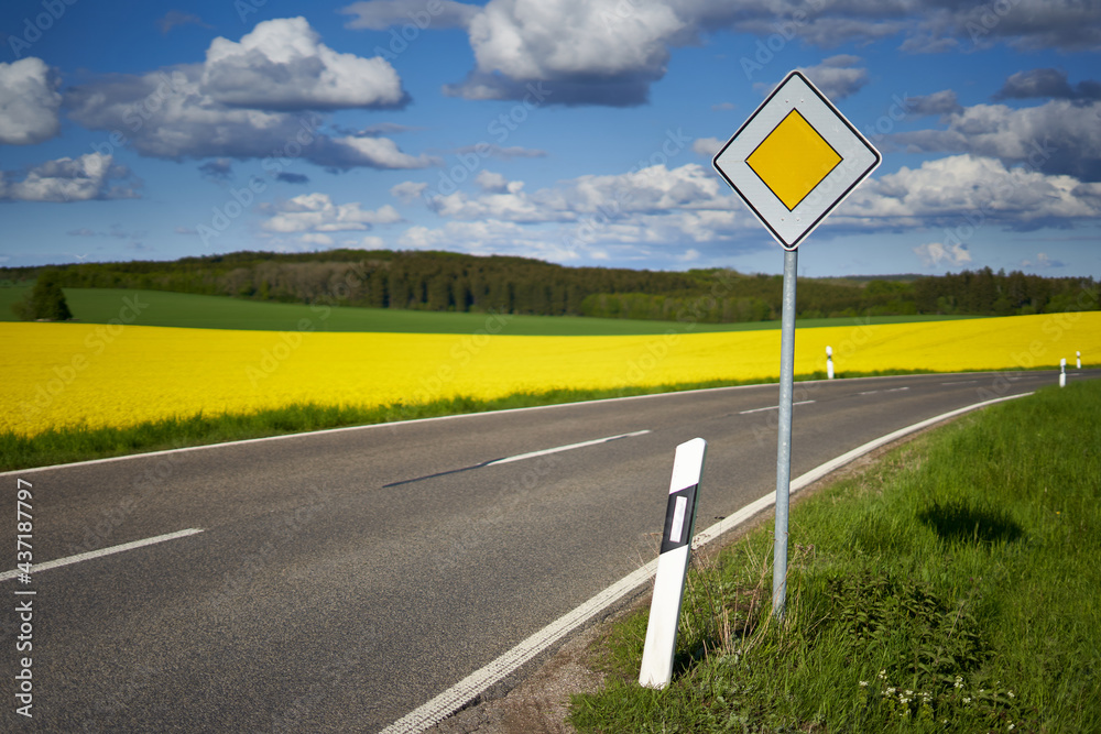 Traffic sign on the side of the road. Yellow rapeseed (Brassica napus) field behind the roadway. Green forest in front of blue cloudy sky. Traffic sign on the side of the road. Yellow rapeseed (Brassica napus) field behind the roadway. Green forest in front of blue cloudy sky.