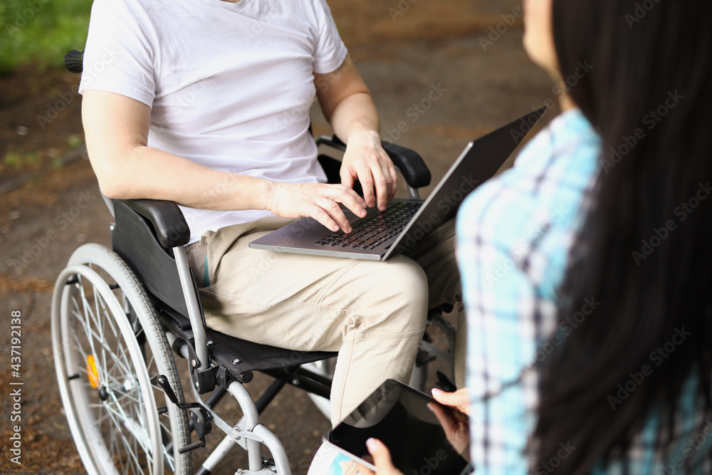 Obraz premium Man in wheelchair works on laptop on street next to woman sits and holds tablet