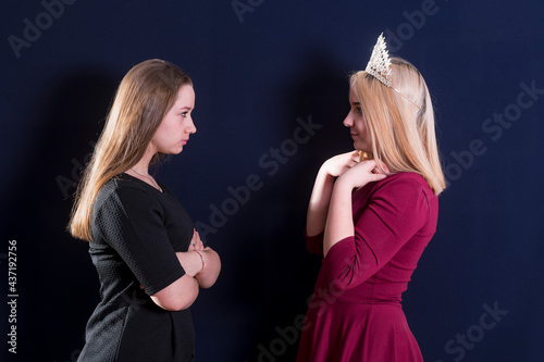 Two young girls, a blonde and a brunette, stand opposite each other. Staring eye to eye. Confrontation. One is wearing a royal diadem.