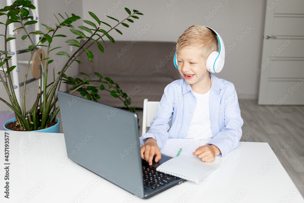 Smiling blonde boy wearing headphones makes a video call via laptop during electronic distance learning at home, concept back to school Smiling blonde boy wearing headphones makes a video call via laptop during electronic distance learning at home, concept back to school