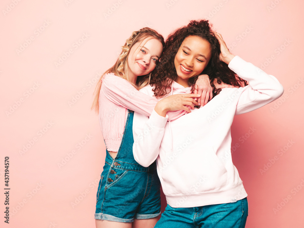 Two young beautiful smiling international hipster female in trendy summer clothes. Sexy carefree women posing near pink wall in studio. Positive models having fun. Concept of friendship Two young beautiful smiling international hipster female in trendy summer clothes. Sexy carefree women posing near pink wall in studio. Positive models having fun. Concept of friendship