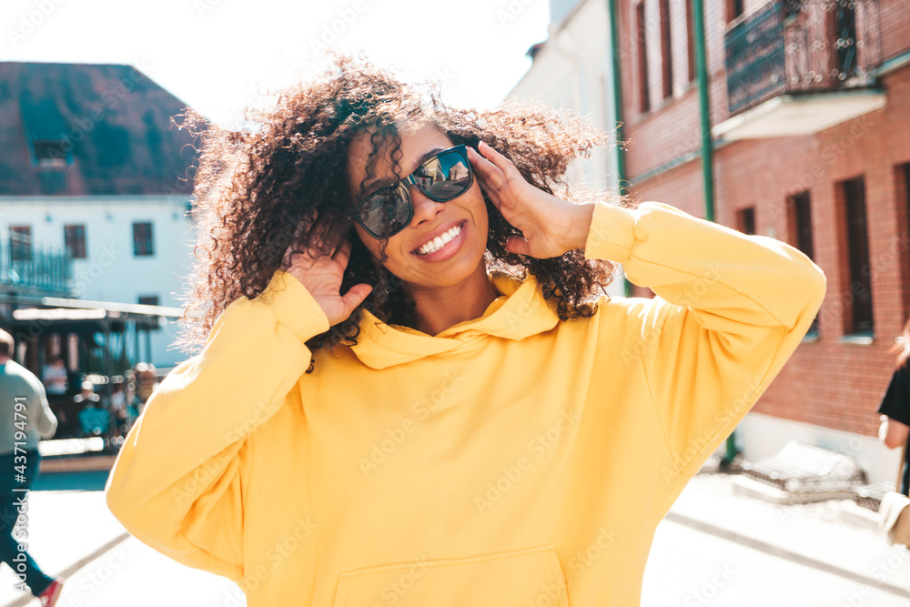 Beautiful black woman with afro curls hairstyle.Smiling hipster model in yellow hoodie. Sexy carefree female posing on the street background in sunglasses. Cheerful and happy