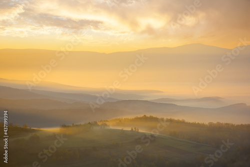 mountainous countryside landscape at foggy sunrise. wonderful autumnal nature scenery with distant rural valley in glowing mist. trees and fields on hills in morning light