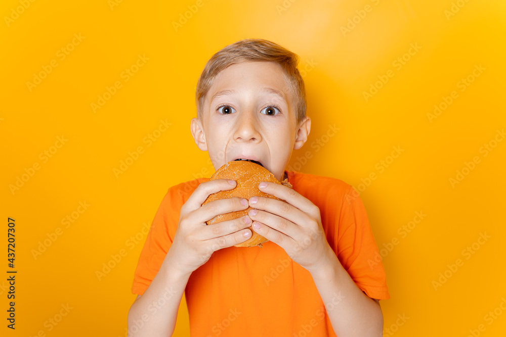 a boy in an orange T-shirt holds a hamburger in both hands and greedily bites it on a yellow background