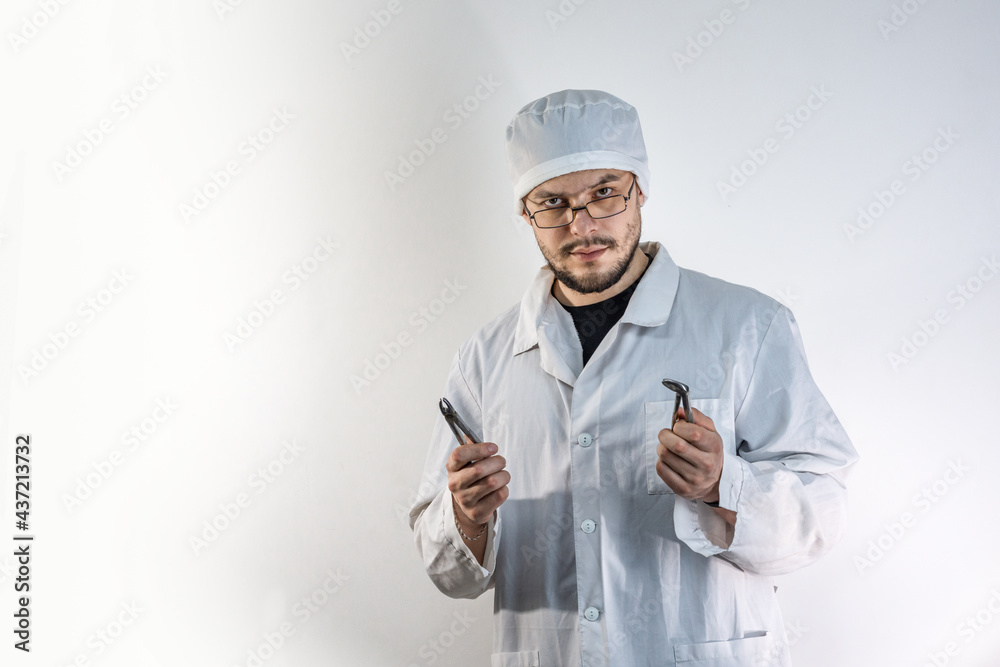 A young, dark-haired man with a beard, wearing a white medical coat and a cap, holds a pair of dental forceps against a light background. A young, dark-haired man with a beard, wearing a white medical coat and a cap, holds a pair of dental forceps against a light background.