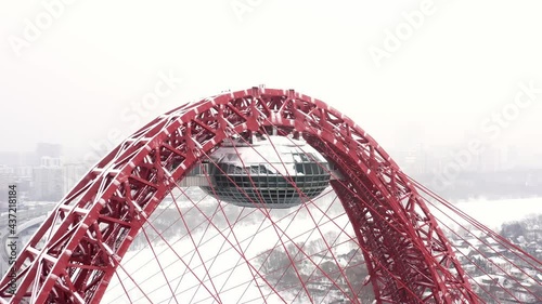 Zoom out drone shot of red bridge over the river with busy traffic, buildings and grey sky at winter time. Zhivopisny bridge in Moscow. Snow photo