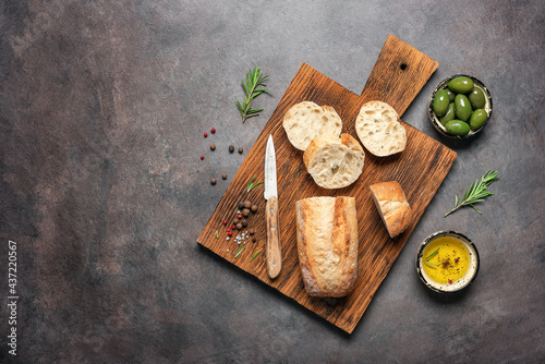 Fresh Italian ciabatta bread on a wooden cutting board with olive oil, olives and herbs, dark rustic background. Top view, flat lay, copy space.