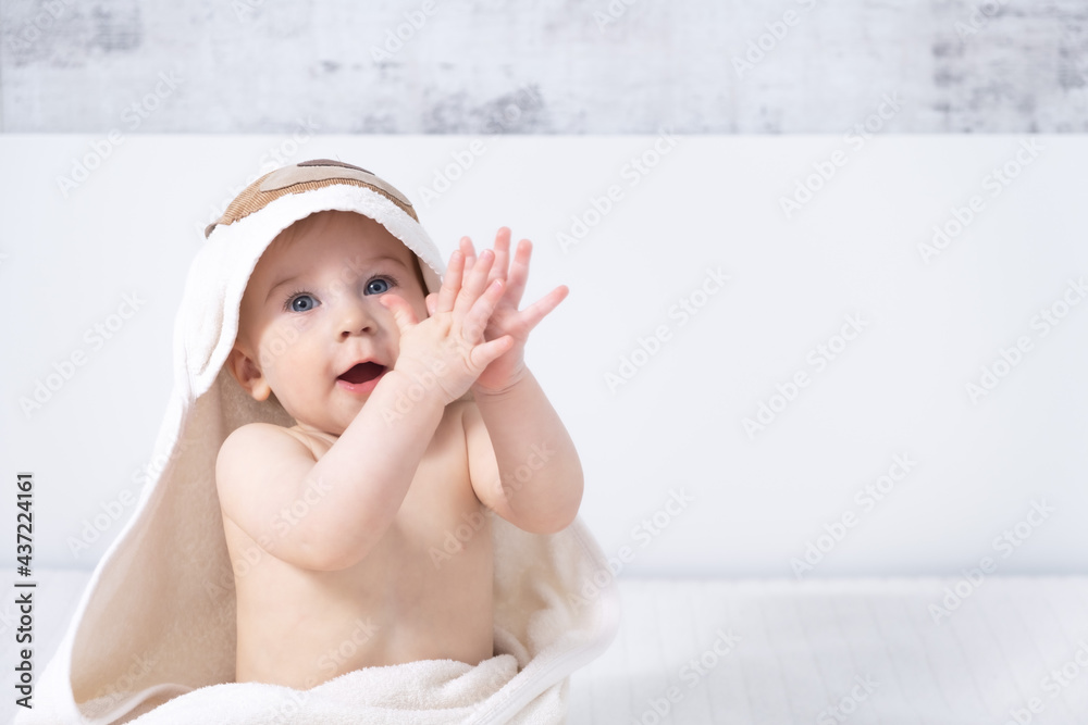 Beautiful ten month happy baby girl sitting on bed claping hands indoors in light modern interior with towel after bath. Baby care concept, copy space Beautiful ten month happy baby girl sitting on bed claping hands indoors in light modern interior with towel after bath. Baby care concept, copy space