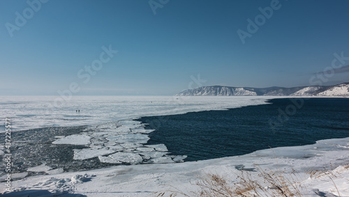 The border between a frozen lake and the source of a non-freezing river. White ice, blue water, melted ice floes. Snow-capped mountain range against the backdrop of the azure sky. Baikal and Angara