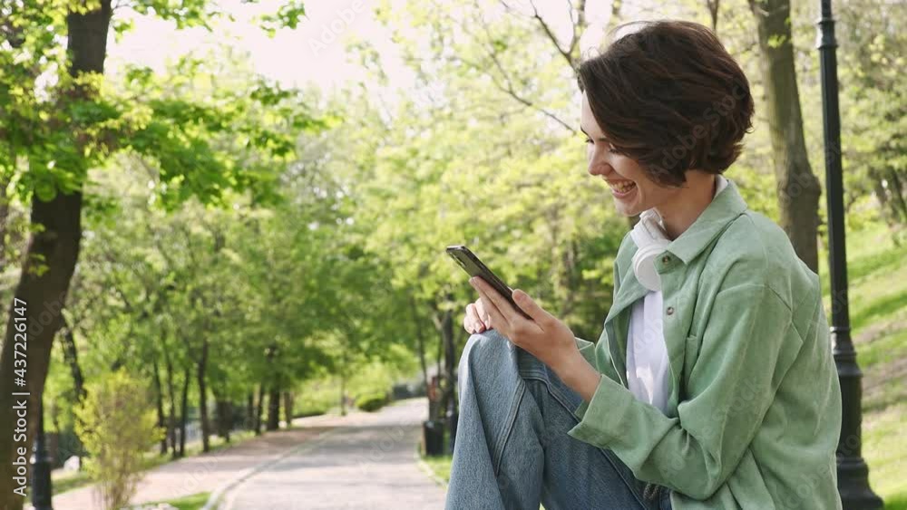 Young woman 20s wearing mint shirt white t-shirt sit on bench hold in hand use mobile cell phone rest relax in spring green city park sunshine lawn outdoors on nature. Urban lifestyle leisure concept