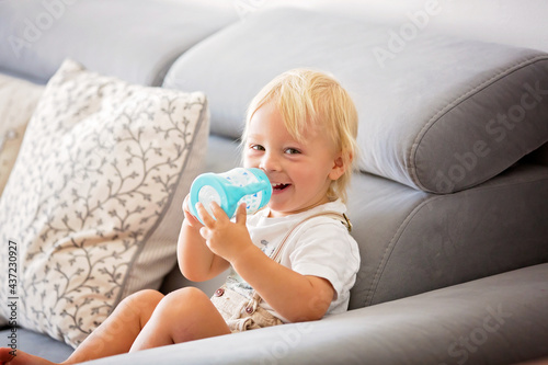Adorable baby boy drinking milk from a bottle in a white sunny living room.