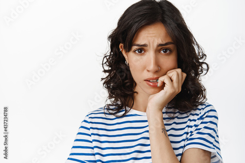 Close up of anxious brunette girl, biting fingernails and looking embarrassed at camera, feel nervous and scared, standing over white background © Cookie Studio