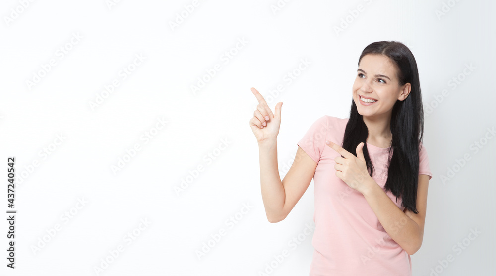 Young woman with white perfect smile in summer clothing shows points fore fingers aside on empty space over white wall. Young woman with white perfect smile in summer clothing shows points fore fingers aside on empty space over white wall.