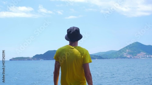 Young caucasian man in yellow t-shirt and black hat looks to the sea and mountains