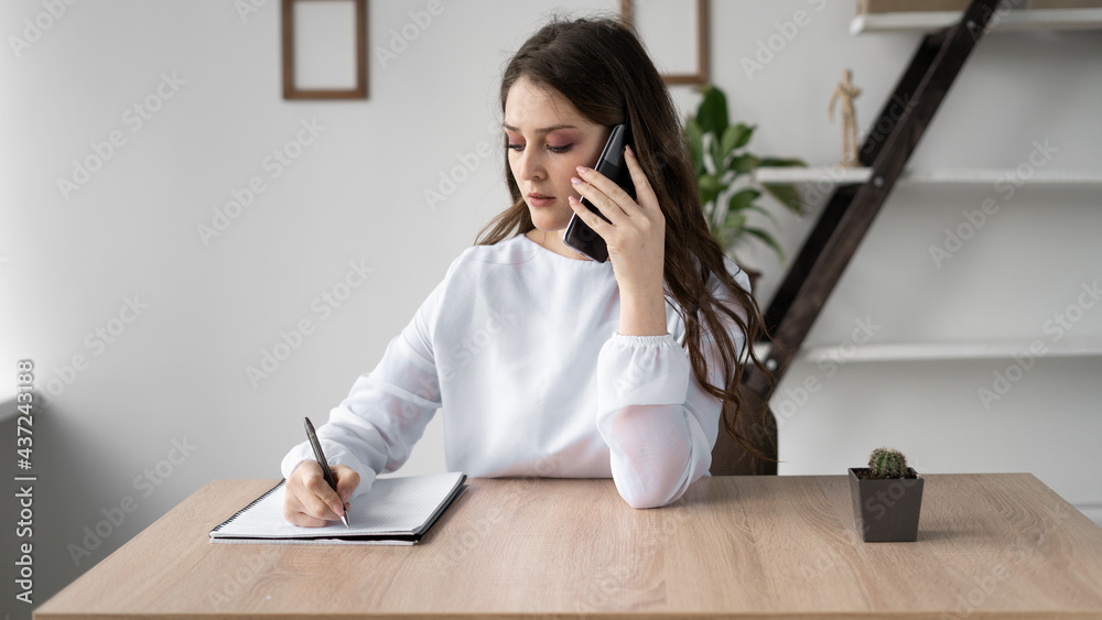 Portrait of a business woman working from home sitting at a desk talking or talking on the phone with a client and making notes in a notebook. Mobile network consulting. office work. Portrait of a business woman working from home sitting at a desk talking or talking on the phone with a client and making notes in a notebook. Mobile network consulting. office work.