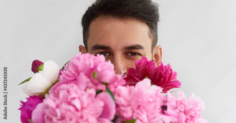 Closeup portrait of man's handsome eyes looking directly to the camera through the huge bouquet of pink peonies as a gift for Valentine's day or wedding day. Smiling male carrying flowers in the hands Closeup portrait of man's handsome eyes looking directly to the camera through the huge bouquet of pink peonies as a gift for Valentine's day or wedding day. Smiling male carrying flowers in the hands