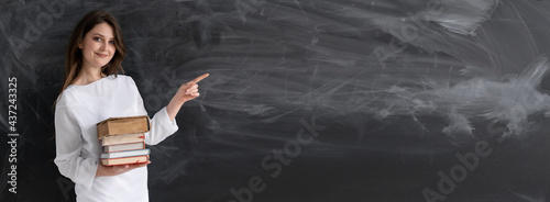 Modern teacher woman 30 years old with textbooks in her hands stands in the classroom near the blackboard with a place for the text. Online lesson and teacher work. School education. Banner. photo