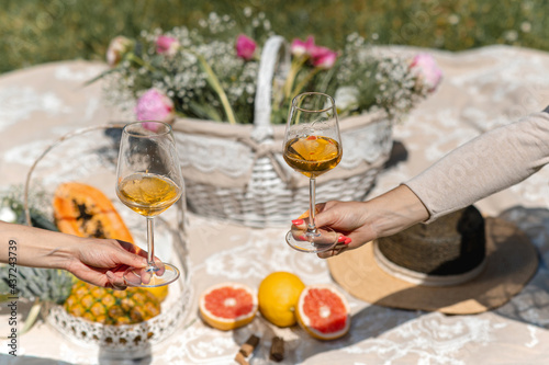Unrecognizable females extending arms to show two glasses with white wine. Blanket picnic on background setting with tropical fruits and flowers.