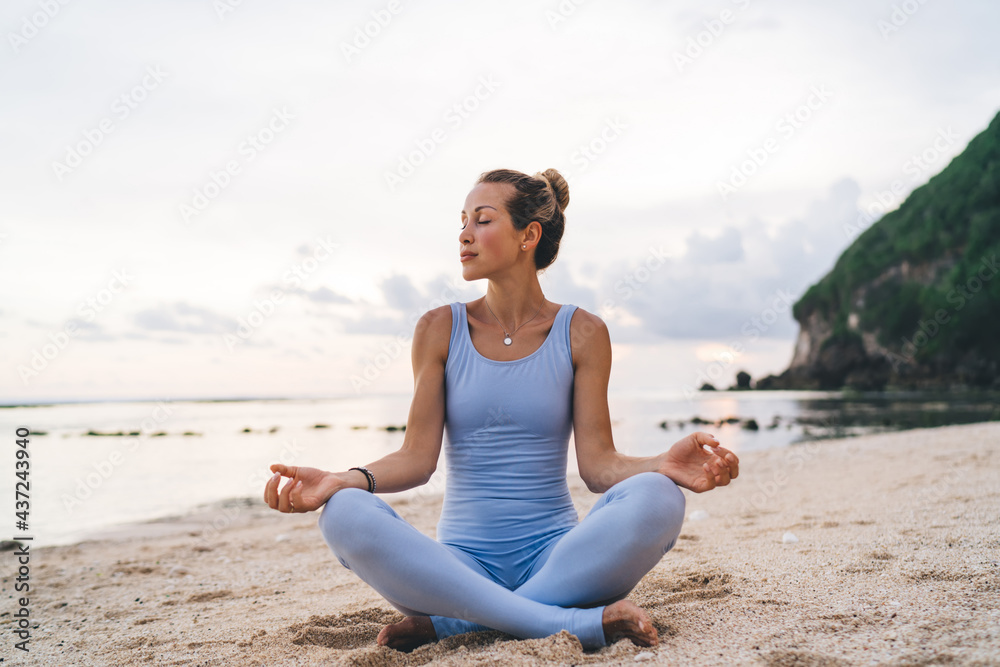 Tranquil female with closed eyes practicing yoga at seashore beach reaching healthy lifestyle and wellness, calm woman with flexible body sitting in lotus pose enjoying asana time for mindfulness
