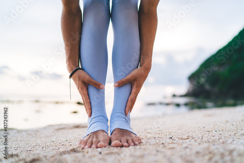 Selective focus on cropped female legs and hands during sportive workout training at seashore coastline, flexible athletic woman stretching body muscles during active physical practice at beach
