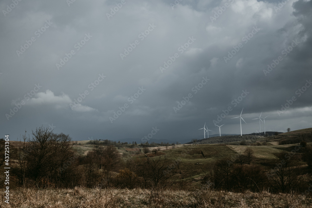 Farm of wind turbines for renewable wind energy on far horizon on hills of Romania under the dark cloudy sky. Copy space. Farm of wind turbines for renewable wind energy on far horizon on hills of Romania under the dark cloudy sky. Copy space.