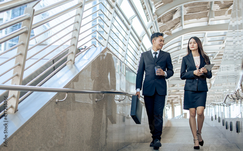 Adult two Asian smart professional businesspeople wearing formal suits, holding cup of coffee and tablet, discussing and talking together with happiness while walking outside office in morning.